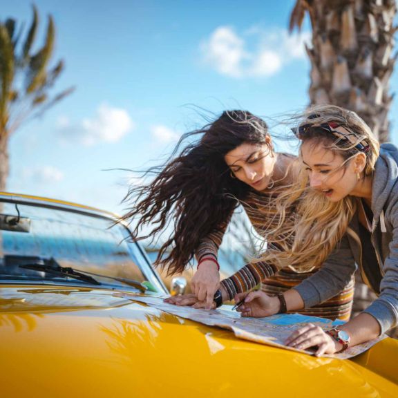 Zwei junge Frauen breiten eine Landkarte auf der Motorhaube aus.