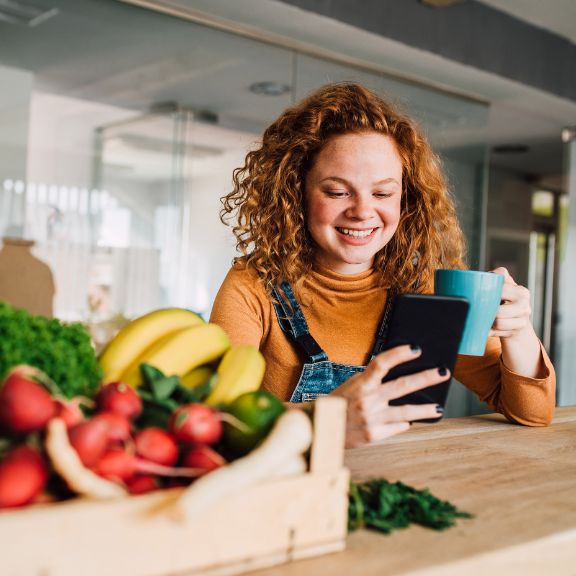 Frau mit Tasse in der Hand und großem Obstkorb neben sich, schaut auf ihr Handy.