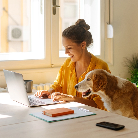 Frau mit Hund arbeitet am Laptop am Schreibtisch, helles Büro mit Fenster und Pflanzen im Hintergrund.