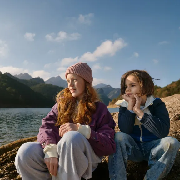 Zwei Kinder sitzen an einem See-Ufer und schauen aufs Wasser.