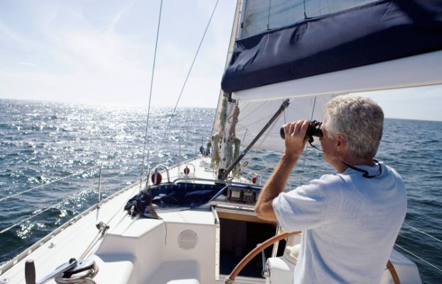 Älterer Mann steuert ein Segelboot und blickt durch ein Fernglas.
