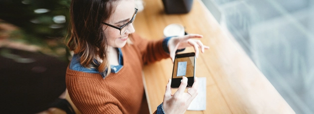Frau mit Brille und orangefarbenem Pullover fotografiert mit Smartphone auf Holztisch