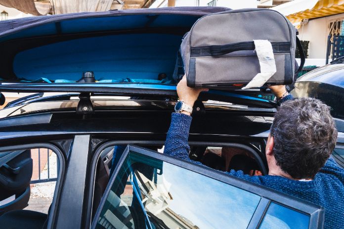 Familienvater mit grauem Haar, der einen Reisetasche in eine Dachbox auf seinem Auto packt.