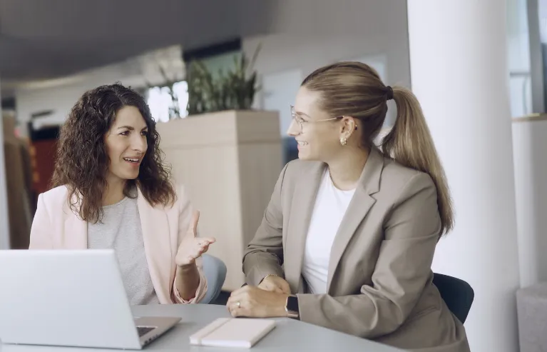 Zwei Kolleginnen unterhalten sich an einem Schreibtisch in einem Büro.
