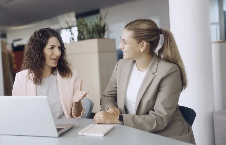 Zwei Kolleginnen unterhalten sich an einem Schreibtisch in einem Büro.
