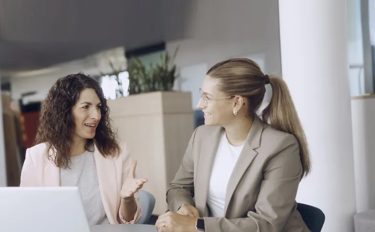 Zwei Kolleginnen unterhalten sich an einem Schreibtisch in einem Büro.