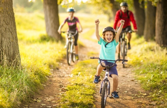 Kind auf Fahrrad fährt freudig auf Waldweg, Familie mit Kindern folgt auf Rädern durch sonnigen Wald.