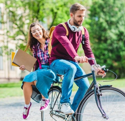 Zwei junge Menschen fahren auf einem Fahrrad durch einen Park, halten Ordner und Hefte, grüne Bäume im Hintergrund.