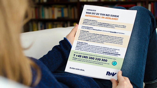 Person in blauem Pullover hält Informationsblatt mit Ratschlägen zu Auslandsaufenthalten und RKK-Kontaktdaten in Wohnzimmer.