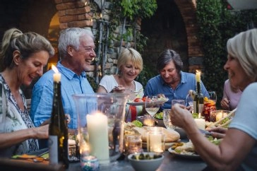 Fünf Personen sitzen beim Abendessen an Tisch mit Kerzen, Flaschen und Speisen unter Backsteingewölbe mit Efeu.