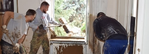 Person reinigt Fensterrahmen mit Bürste, Wald im Hintergrund, alte weiße Fensterbank mit Farbabplatzungen sichtbar.