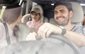 Zwei Personen sitzen lächelnd im Auto, der Fahrer trägt ein gestreiftes Shirt, der Beifahrer eine rosa Kappe und helle Kleidung.
