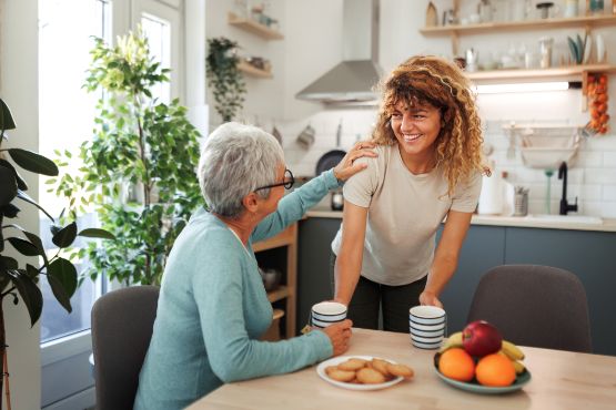 Mutter sitzt mit Kaffeetasse am Esstisch und greift zugewandt die Schulter ihrer am Tisch stehenden Tochter.er 
