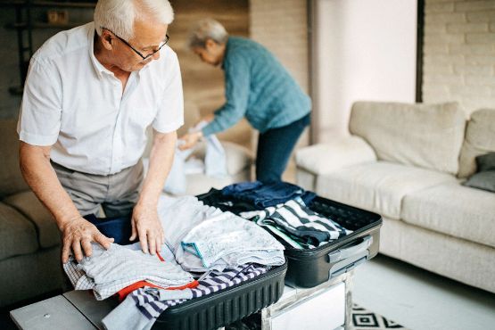 Aelterer Mann packt Koffer für die Klinik mit seiner Frau im Hintergrund