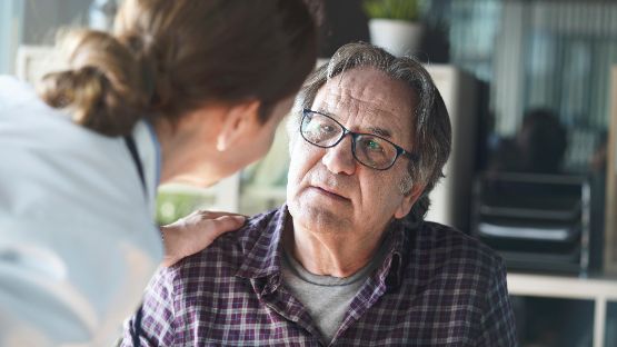 Weibliche Pflegefachperson beugt sich zu pflegebeduerftigem aelterem Mann Hand auf die Schulter legend