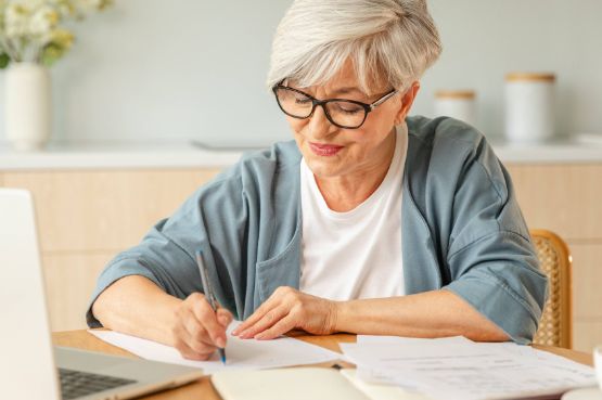Frau macht sich Notizen auf Papier am Küchentisch mit Laptop vor sich
