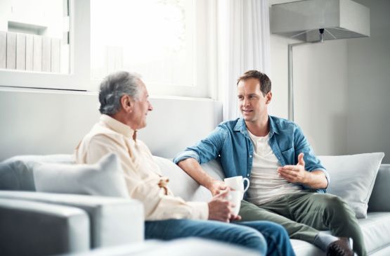 Pflege durch Angehörige: Erwachsener Sohn und älterer Vater auf dem Sofa.