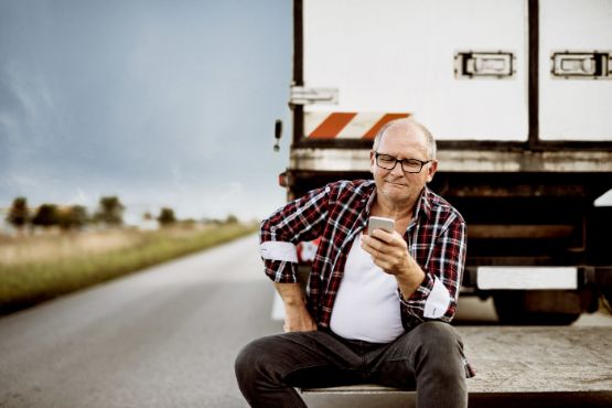 Truck-Fahrer sitzt auf der Laderampe und schaut auf sein Smartphone.