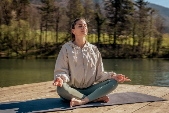 Frau im Trainingsoutfit sitzt am See und praktiziert Yoga.