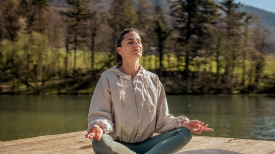 Frau im Trainingsoutfit sitzt am See und praktiziert Yoga.