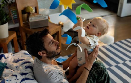 Ein Vater hält sein Baby hoch, damit es mit einem Mobile spielen kann, als Symbol für finanzielle Absicherung.