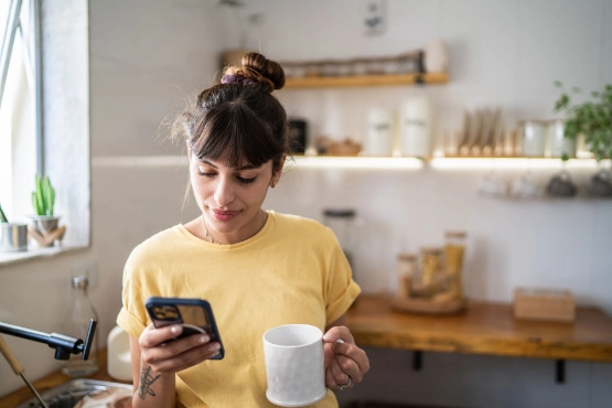 Eine Frau schaut auf ihr Handy, in der anderen Hand hält sie eine Kaffeetasse.