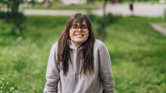 Junge Frau mit langen dunklen Haaren und Brille sitzt im Park auf einer Bank und lächelt in die Kamera