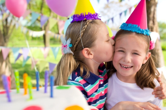 Zwei Mädchen mit Partyhüten küssen sich lachend auf einer bunten Geburtstagsfeier im Freien.