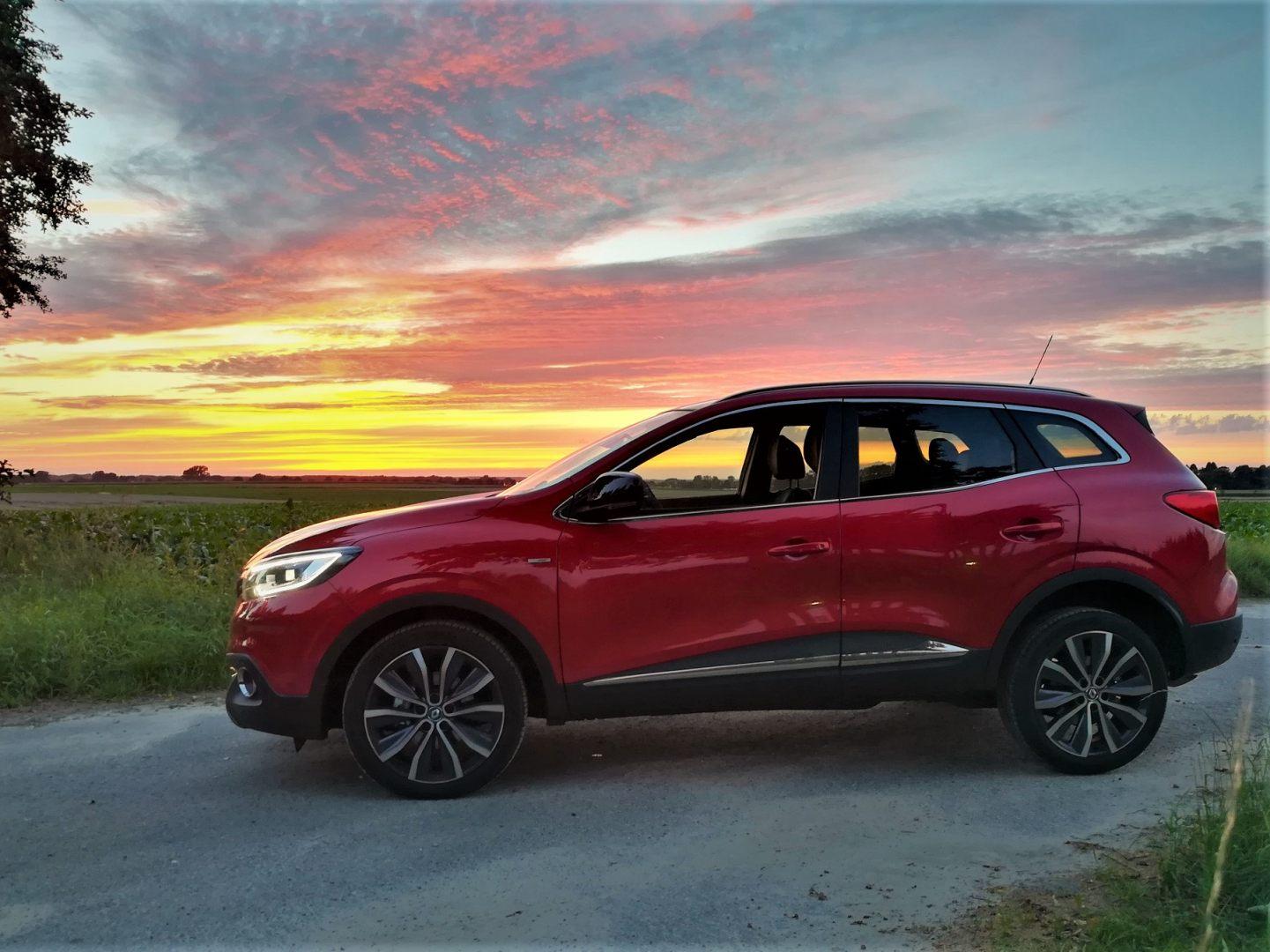 Roter SUV parkt bei Sonnenuntergang auf Landstraße mit dramatischem Himmel und Grünland