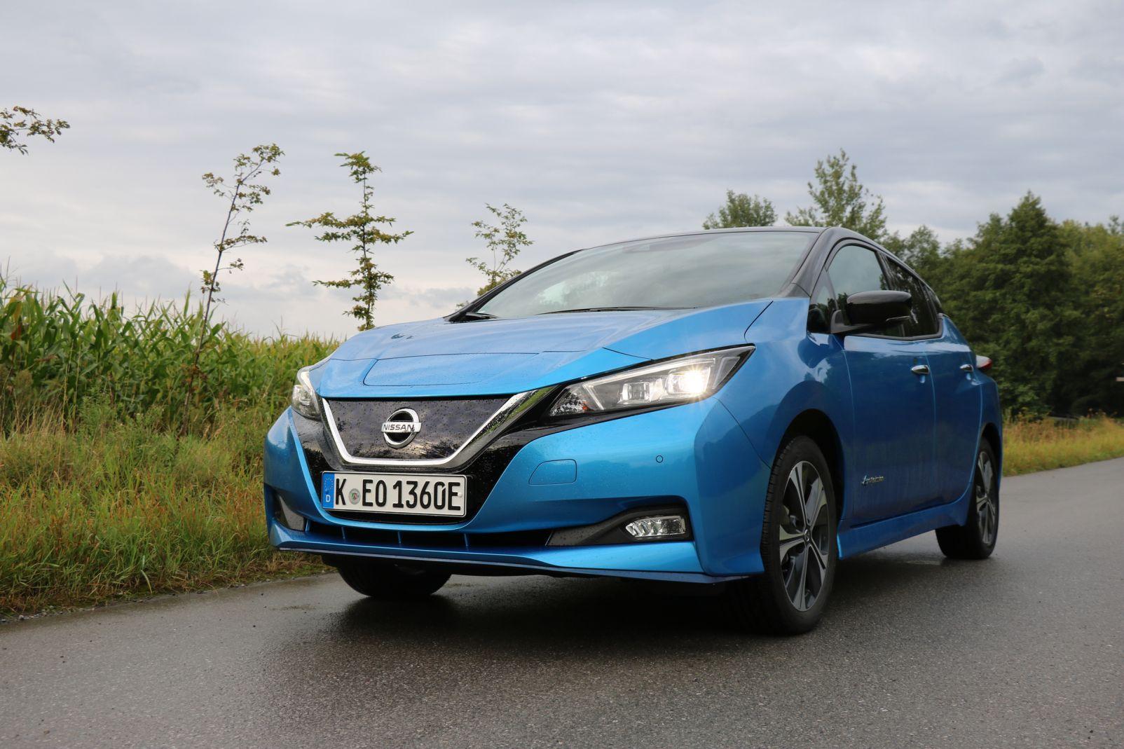Blauer Nissan Leaf Elektroauto auf Landstraße neben Maisfeld und Bäumen, bewölkter Himmel