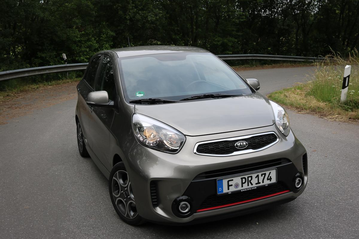 Silberner Kia Picanto Elektroauto auf Landstraße mit Metallleitplanke und bewaldeter Umgebung, Kennzeichen sichtbar