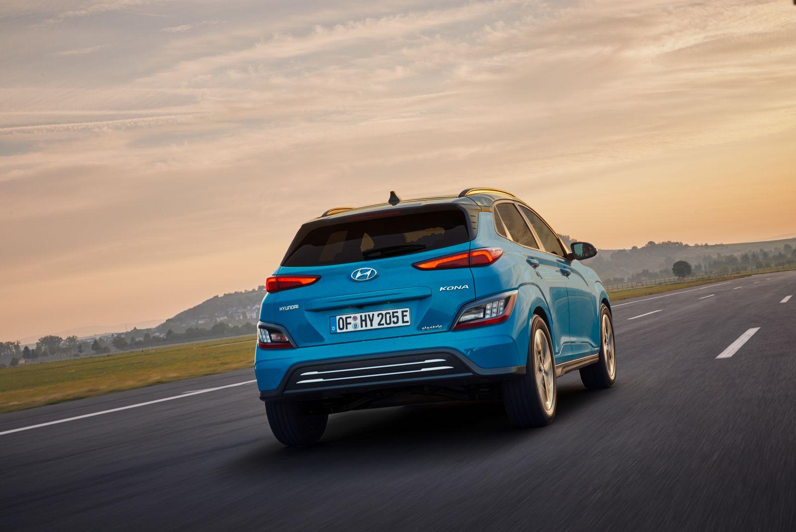 Blauer Hyundai Kona Elektro fährt auf Autobahn, Landschaft mit Hügeln im Hintergrund, sonniger Himmel